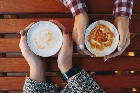 Female hands holding cups of coffee on wooden table background. Big mugs of hot latte drink warming woman hands with freckles. Coffee latte in coffee shop with copy space.の写真素材
