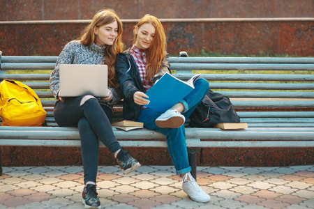 Attractive Young Adult Female Students on Bench Outdoors with Books and Laptop. College Students Internet Computer Sitting Bench Outside Campus Autumn Looking in a Book. Teen Girls Studying.の写真素材