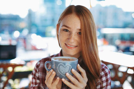 Beautiful woman enjoying the coffee aroma while holding the cup with hands looking to camera....