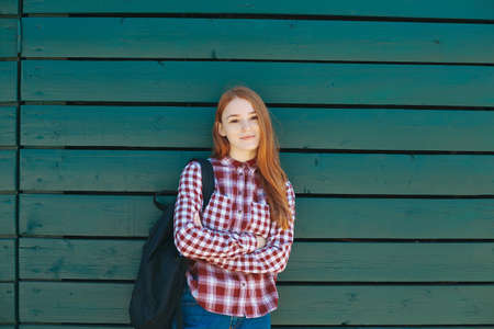 Handsome student with backpack standing outside on green wooden background. Portrait of reddish high school girl dressed in plaid shirt and jeans smiling holding crossed hands, copy space available.の写真素材