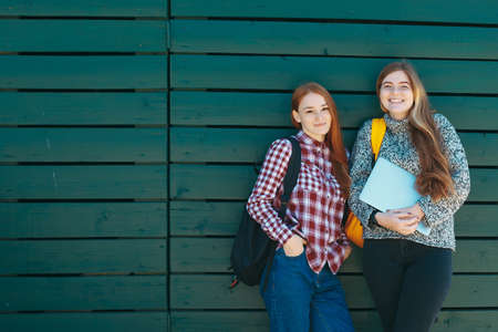 Two college students or high school girls on green wooden background smiling with backpacks and laptop in hands. Students Youth Friendship Concept. Teenagers Young Team Together Cheerful.の写真素材