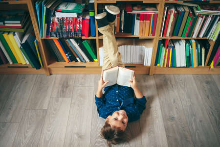 Top view, boy laying on the floor with the feet up, reading a book against multi-colored bookshelf in library.の写真素材