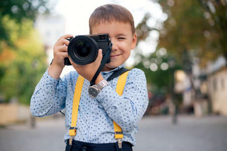 Boy, in blue shirt, silver watch and yellow suspenders, makes pictures on film retro style digital camera, stylishly dressed, photographer, outdoor portrait, close up, street photo.の写真素材