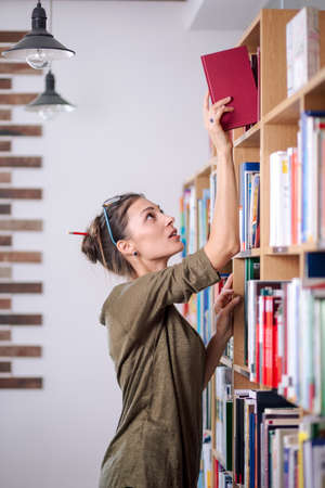 Young attractive woman wearing glasses and cotton blouse looking for a book on a colorful bookshelf,...