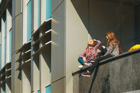 College students take a break after classes. Young girls with reddish hair sit on a modern building near windows on campus outdoors.の写真素材