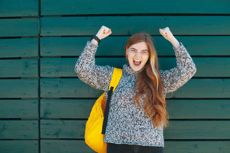 Portrait of excited smiling college student holding hands up and opened mouth standing on green...