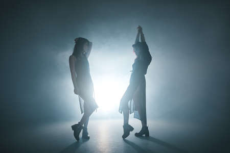 Stylish girls having fun, posing, dancing with the light play and fume. Studio shot of two gorgeous ladies moving in smother with illumination from behind, creating illusions and fantasies.の写真素材
