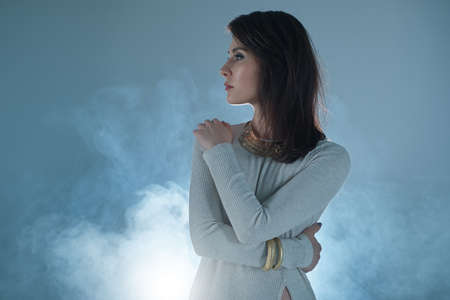 Beautiful young woman profile portrait over smoky background hugging herself, dressed in white...