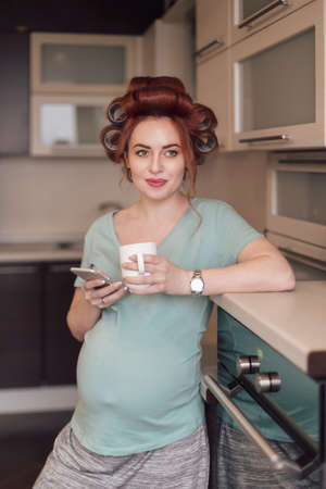 Cheerful, smiling, pregnant woman using a smartphone, looking to screen, to camera while holding a cup of tea or coffee in the kitchen. Female searching facts about pregnancy.の写真素材
