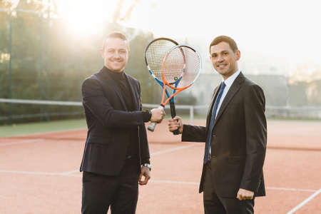 Two businessmen in suits holding tennis rackets crossed looking to camera, announcing their intention to win, smiling being ready to compete. Business, sport, competition, partners.の写真素材