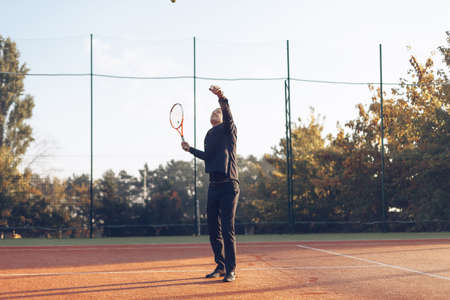 Portrait of confident businessman holding tennis racket serving against tennis court background. A...