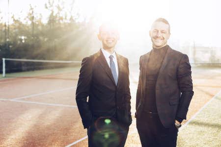 Two handsome young businessmen in classic clothes are looking at camera and smiling, standing in sunlight with hands in pockets outdoors. Business and sport. Fresh mornings in business.の写真素材