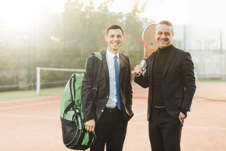 Business, partnership and sport concept. Portrait of smiling successful business men standing in friendship pose holding tennis racket and bag on shoulder, on a tennis court background.の写真素材