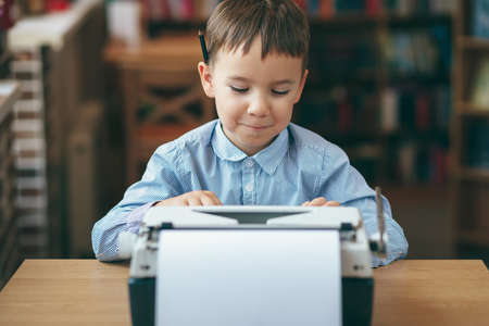 Smiling preschool journalist boy sitting at table and typing typewriter with  a pencil on the top of year. Businessman using typewriter at desk in office. Business and new technology.の写真素材