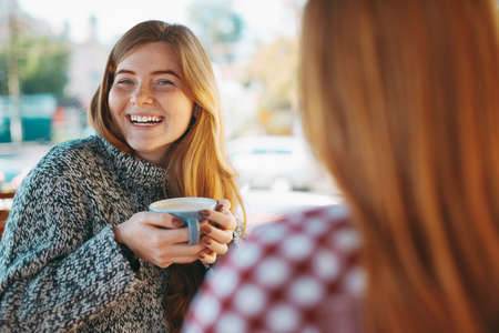 People, communication and friendship concept - laughing young women having fun while drinking coffee and chatting at outdoor cafe. Two natural reddish girls having coffee.の写真素材