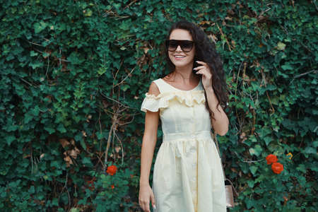 Close-up portrait of beautiful caucasian woman with charming smile walking, posing outdoors, on plants background.  Model wearing stylish sunglasses.の写真素材
