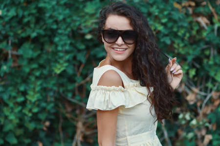 Close-up portrait of beautiful caucasian woman with charming smile walking, posing outdoors, on plants background.  Model wearing stylish sunglasses.の写真素材