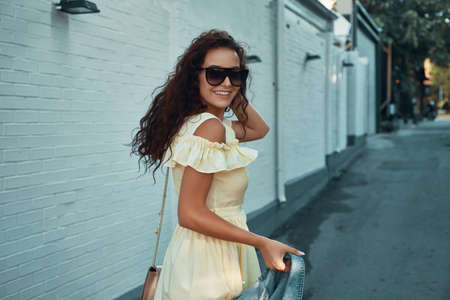 Portrait of inspired lady spending vacation in Europe. Outdoor photo of smiling, cheerful girl with brown curly hair, sunglasses, light yellow summer dress, walking around city in the morning.の写真素材