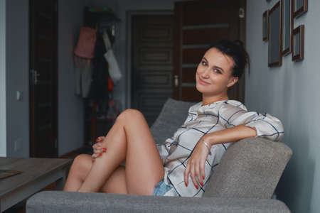 Portrait of beautiful smiling young woman sitting on sofa. Attractive female relaxing at home after...