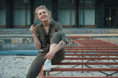 Portrait of a casual blond girl sitting on the metal construction with a building in the background. Young modern woman posing in the city, outdoors. Urban lifeの写真素材