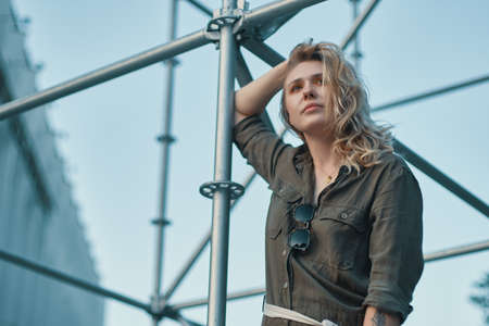 Fashion shot of carefree young woman in stylish casual romper hanging on a silver metal railing construction with a building, stairs, sky in bkgroundの写真素材