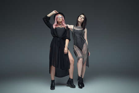Two fashionable girls wearing black trendy outfits, accessories, sunglasses, hat and leather boots posing on studio gray background standing in fashion body pose.の写真素材