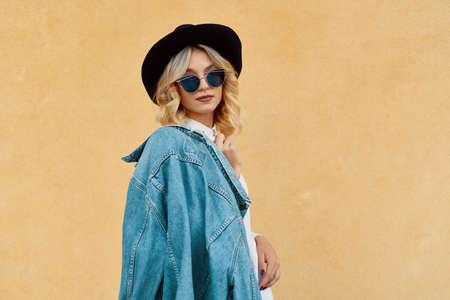 Portrait of Hipster Fashion Girl Holding Jaket on one Shoulder Standing at the Yellow Wall Background. Youth Urban Fashion Concept. Copy Space.の写真素材