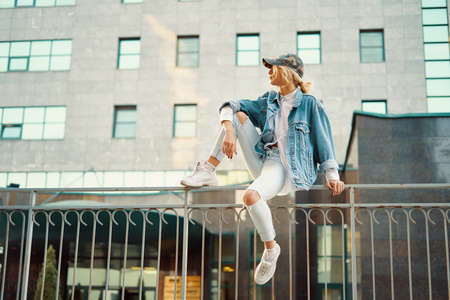 Pretty girl sits on the fence railing in a European city with a modern building in the background...