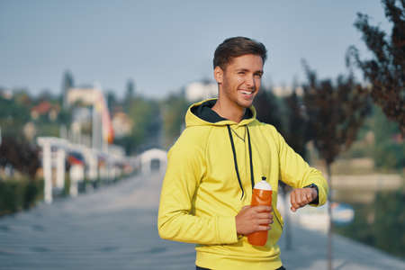 Portrait of handsome young man wearing sportswear checking smart watch while running outdoors in...