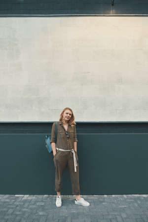 A hipster girl with long blonde hair, wearing a stylish romper and a backpack is looking aside while standing on a wall background on a street. Horizontal mock up. Empty space for text or design.の写真素材