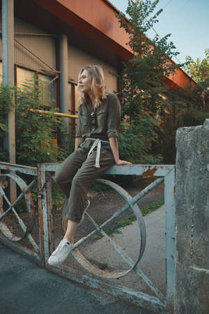 Stylish girl posing in the street sitting on a steel fence with bent legs. Urban style. Street fashion.の写真素材