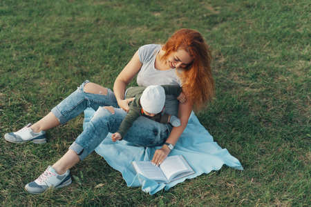 Beautiful mom is reading a book with her cute baby boy and smiling, while sitting on the grass