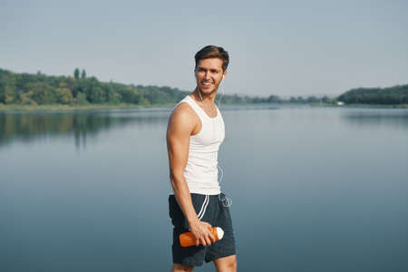 Portrait of smiling and relaxing athletic man holding bottle of water in his hands. Sportsman in black sportswear wearing white earphones enjoying morning while jogging near a lake in the neighborhoodの写真素材