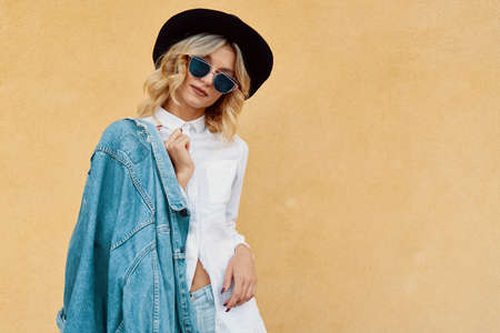 Portrait of Hipster Fashion Girl Holding Jaket on one Shoulder Standing at the Yellow Wall Background. Youth Urban Fashion Concept. Copy Space.の写真素材
