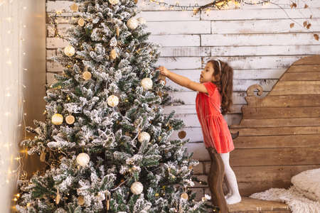Merry Christmas and Happy Holidays. Cute little girl is decorating the Christmas tree holding a silver ball in her hands, stretching to the top while standing on a wooden chair.の写真素材