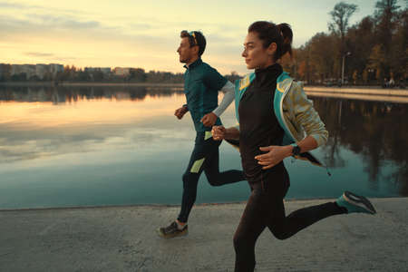 Motivated couple of runners out for a run on the lake at the sunrise. Young man and woman in sport clothing, running together. Sportive people training outdoorsの写真素材