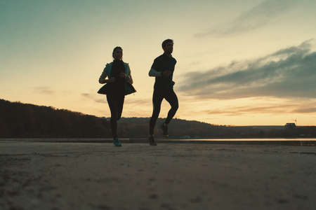 Motivated couple of runners out for a run on the lake at the sunrise. Young man and woman in sport clothing, running together. Sportive people training outdoorsの写真素材