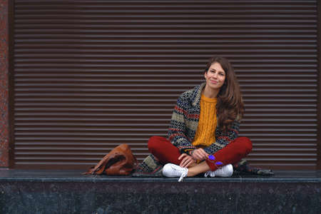 Portrait of young smiling woman sitting crossed legs, posing in street of a European city. Braun copy space in background on the rightの写真素材