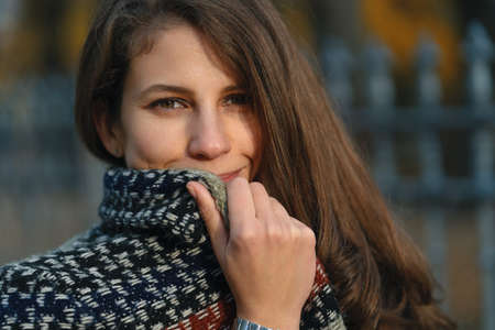 Close-up portrait, woman hiding, smiling face behind coat collar. Natural brown wavy hair and eyes,...