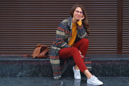 Portrait of young, beautiful woman wearing a stylish coat, red pants, orange sweater, white sneakers, with a brown leather bag, posing in street of a European city. Copy spaceの写真素材