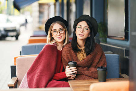 Autumn winter portrait of two young women in an outdoor cafe, drinking coffee to go, talking. Urban background. Sistersの写真素材