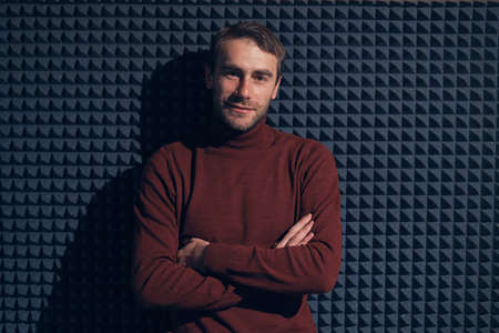 Young, handsome man leaning against grey wall with arms crossed. Smiling male looking at camera. Portrait of a confident young man standing over grey background with geometric forms and copy space.の写真素材