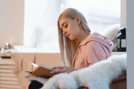 Young woman chilling at home in a comfortable round armchair near the window. Girl relaxing, reading and dreaming book in the loft living room. Imagining the book extraordinary world.の写真素材