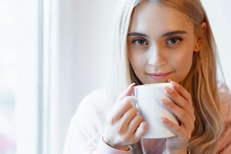 A pretty smiling girl enjoying a cup of coffee or milk while sitting by the blurred window glass. Wearing a casual pink, pale parka. Close up white mug in woman hands near lips.の写真素材