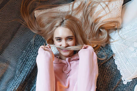 Carefree beautiful girl playing with her long blond hair while lying on the bed at home. Happy weekends, having fun while enjoying and relaxing. Top view.の写真素材