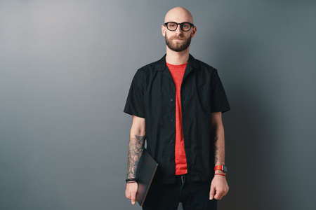 A Young bearded man with glasses holding laptop under the arm in the studio on gray background....