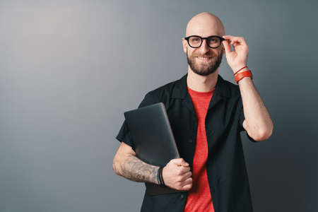 Smiling young bearded man holding laptop under the arm touching  his glasses with fingers, in the...