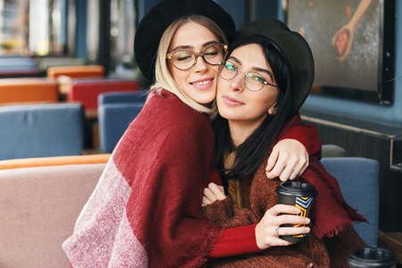 Autumn winter portrait of two young women in an outdoor cafe, hugging, drinking coffee to go. Urban...
