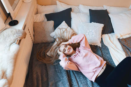 Beautiful smiling girl, with long blond hair, enjoying happiness, she cant belive its true. Portrait gorgeous young woman lying on the bad with pillows in comfy pink pale parka looking at camera.の写真素材