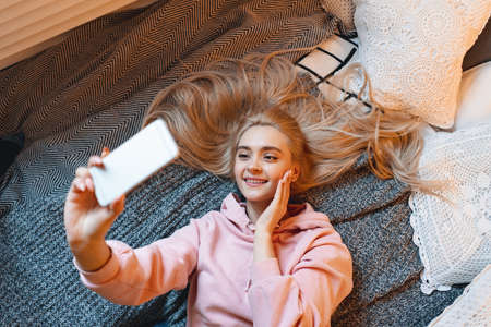 Pretty young girl having fun, making selfie while lying on the bed at home. Top angle view. Charming Caucasian woman taking picture of herself, for her friends and fans, for social media.の写真素材
